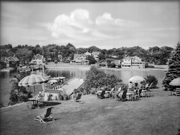 Elevated view of tables, chairs, and umbrellas arranged on a hotel lawn near a boat dock. People mill about on the dock, and two women are sitting beneath an umbrella on the right. Numerous houses stand along the shore on the opposite side of the harbor.