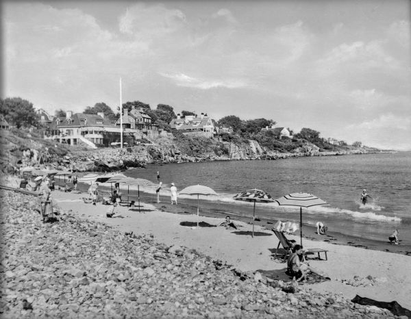 View toward bathing beach and seashore, with people sunbathing near umbrellas and wading in the water. Summer homes are in the background on the cliffs along the seashore.