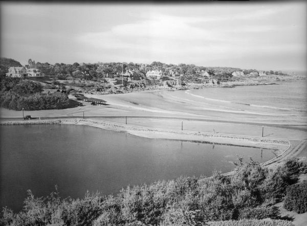 Elevated view of a pond in the foreground, and the seashore beyond. Houses are along the cliffs in the background.