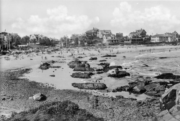 View along rocky shoreline towards adults and children on the seashore, nde wading in the water. In the distance are cottages and other buildings, possibly summer homes and hotels.