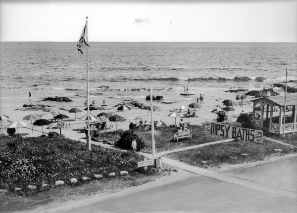 Elevated view of the Dipsy Baths outbuilding and surrounding beach. Umbrellas are set up in the sand where people sunbathe or wade in the water, and an American flag flies from a pole in the grass.