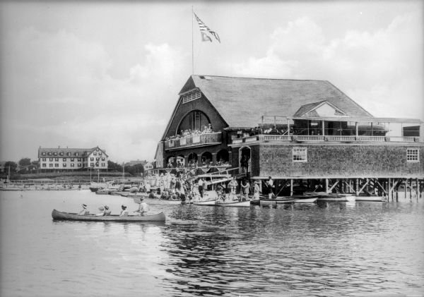 View across water toward a crowd of people standing on both the upper and lower level floors of a boathouse overlooking the lake. On the water beside the structure are numerous canoes and rowboats, many filled with small groups of people.