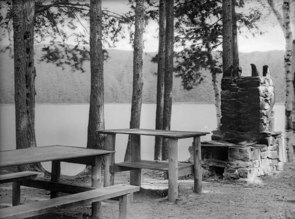 A stone fireplace and wooden tables are set up along the shore at Wabanaki Lodge. The area is surrounded by trees and overlooks a heavily-wooded area on the opposite shore.