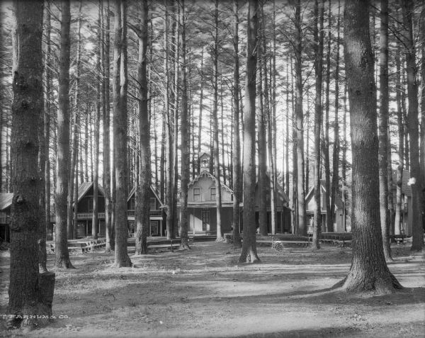 A row of resort cottages as seen through trees in a wooded area. The buildings each have two stories and a gabled roof. The middle structure is a church, as signified by the bell tower and cross.