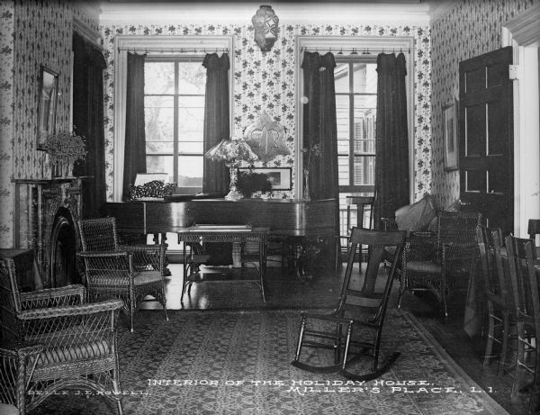 A view of the parlor at Holiday House. A piano stands near the windows and the room is furnished with a seating area focused around a fireplace on the left wall, consisting of wooden and wicker chairs and an area rug. Caption reads: "Interior of the Holiday House, Miller's Place, L.I."