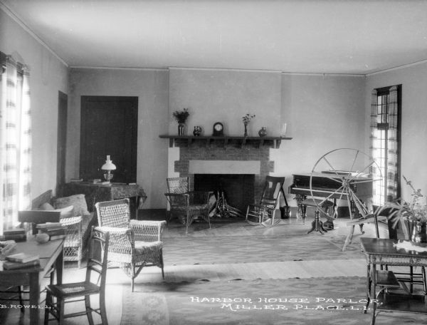 View of the parlor with a settee and a desk that line the outer walls. A spinning wheel and a piano are on the right near a window. Two chairs are in front of the fireplace on the back wall. Caption reads: "Harbor House Parlor, Miller Place, L.I."