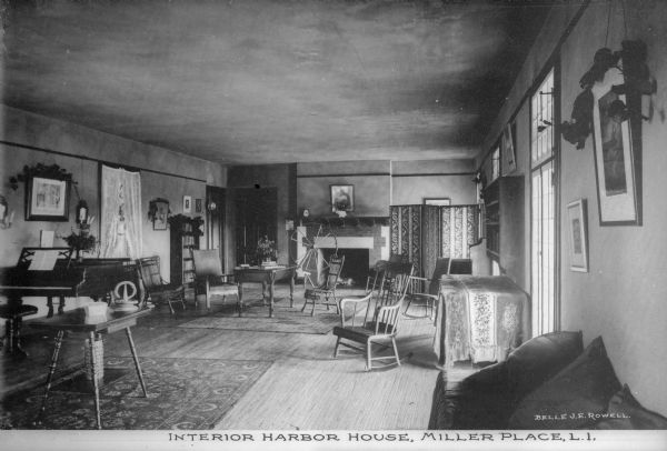 View of the parlor, with tables, chairs, and bookcases, as well as a piano and spinning wheel. A fireplace is on the far wall. Caption reads: "Interior, Harbor House, Miller Place, L.I."