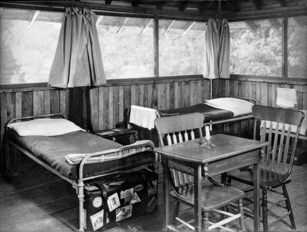 A view of the cabin's interior with two twin-sized beds placed against the wall. The curtains to the windows are open. Suitcases sit at the bottom of each bed, and in the foreground are two chairs at a table with a candle placed upon it.