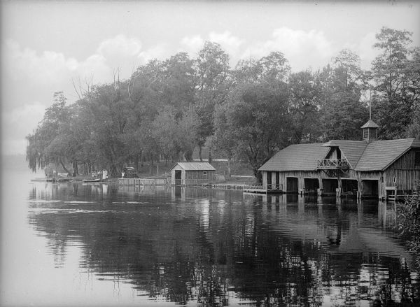 View across water towards two boathouses. The one in the center is small with one door, and the other boathouse on the righthas five doors. To the left of the structures is a group of adults and children.
