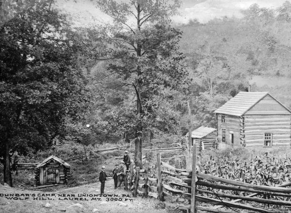 View downhill toward four men standing on the side of the road, with two men leaning against a fence, and one sitting on the fence. To the right is a cornfield and a two-story log house with outbuildings. Caption reads: Dunbar's Camp, near Uniontown, PA. Wolf Hill, Laurel Mt. 3000 Ft."