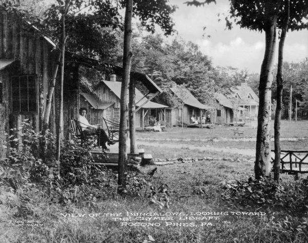A view of a row of wooden vacation homes bordering a wooded area. In the foreground a woman sits on her front porch and looks toward the camera. Others people sit on porches of the homes in the background. Caption reads: "View of the Bungalows, Looking Toward the Clymer Library. Pocono Pines, PA."