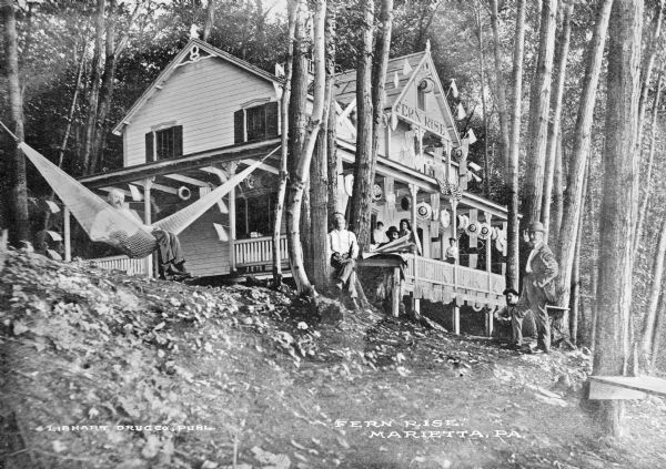 View uphill toward a group of people posing on and near the porch of Fern Rise, possibly a hotel or inn. Decorations and flags hang from the building's porch and roof, and a man sits in a hammock strung between two trees on the left. Caption reads: "Fern Rise, Marietta, PA."