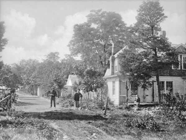 View down dirt road toward a woman sitting on the porch floor of a cottage, while two men stand in the road with a dog lying at their feet. Trees line the street and surround the house, allowing only partial views of the other structures that line a dirt road behind it.