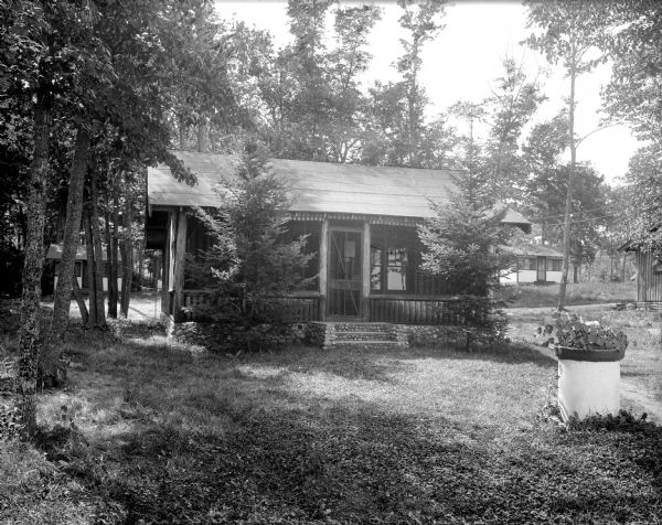 A view of a log cabin at Smoky Point Resort, with two other buildings in the background. The cabin has a stone foundation and a set of steps to the front door. The entry to the structure is shielded by a screened porch and a flower pot stands in the front yard.