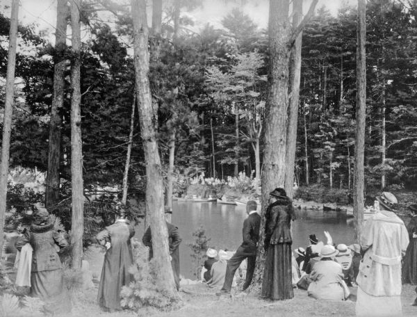 View looking toward a tree-lined shoreline, with men and women watching a canoe race. People are sitting in canoes lining the opposite bank.