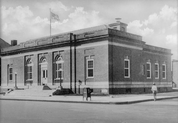 View from intersection toward the facade and right side of the post office. The building consists of brick with stone accents on the top of the arched front windows.
