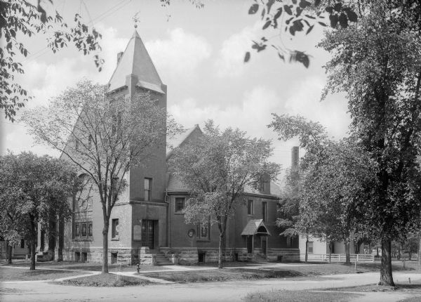 View across street toward the Congregational Church, which features a large stained glass window on the left side of the structure. The building is brick with a stone foundation. A smaller building, possibly a cleric's residence, is on the right.