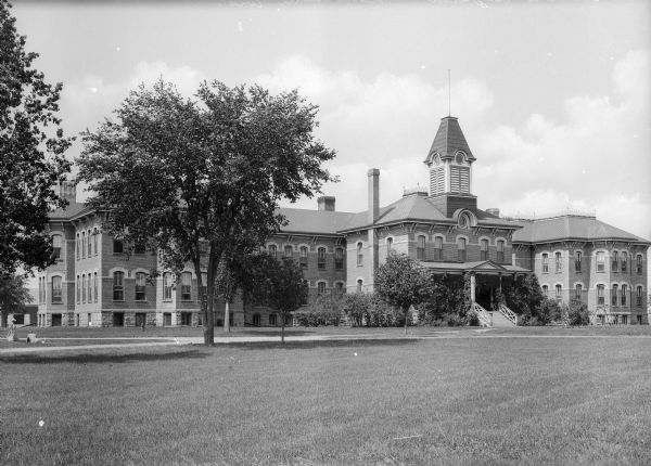 View across lawn toward the stone and brick Italianate-style asylum. The front entrance has a vine-covered porch in front, with bushes and shrubs flanking each side.