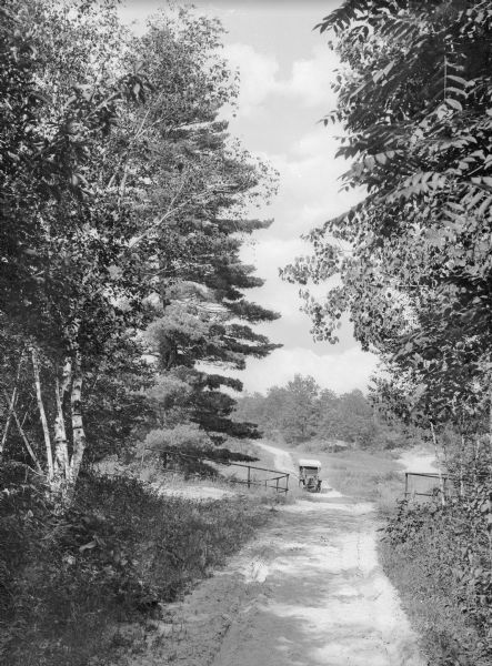 View down unpaved road lined with trees toward an automobile driving up the lane toward a fence.