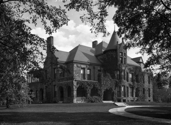 View across lawn toward the facade and right side of the residence of Paul C. Wilson, with a rounded walkway to the right. The stone structure displays some characteristics of the Richardsonian Romanesque style, including large stonework, archways, and heavy massing. The turret and other parts of the facade are covered in vines.