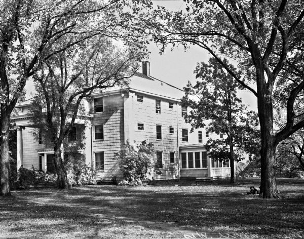 View across yard toward a three-story home surrounded by trees. The pillars of the two-story porch and the corners of the structure resemble Corinthian columns. A man stands against the left pillar. To the right is a woman is reading a book while lying on a blanket.