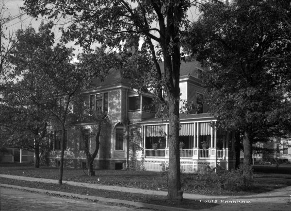 View from street toward a house on a corner. The house has clapboard siding, an asymmetrical roof line, and decorative woodwork on the porch. Striped shades are mounted between the porch columns.