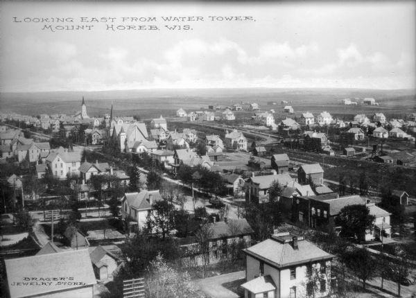 Elevated view of Mount Horeb from the water tower. Homes and commercial structures dominate the left side, while farmland and newly-developed tracts are to the right. Caption reads: "Looking East from Water Tower, Mount Horeb, Wis."