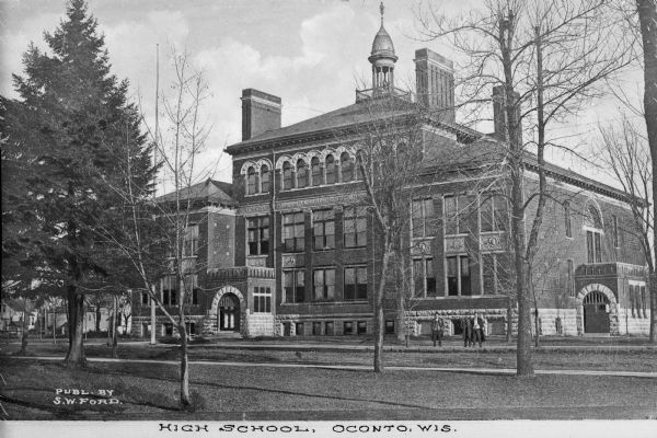 View across lawn toward a high school, with a group of three men standing outside to the right. The school features a dome, arched doorways and windows, and decorative stonework. Caption reads: "High School, Oconto, Wis."