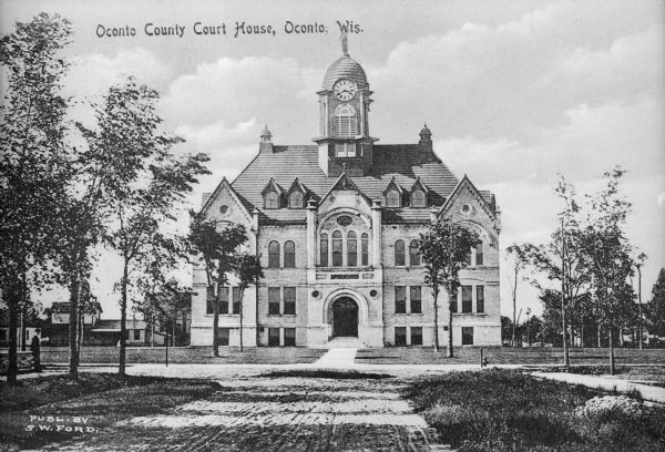 View down unpaved road toward the courthouse facade featuring a domed clock tower. The  structure is made of stone and has rounded windows, steeply-pitched dormers, and an archway above the entrance. Caption reads: "Oconto County Court House, Oconto, Wis."