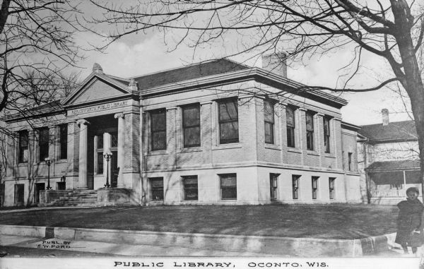 View from street toward the Farnsworth Public Library. The building is constructed of stone and brick; columns and a pediment frame the main entrance. A girl wearing a winter coat is on the sidewalk to the right. Caption reads: "Public Library, Oconto, Wis."