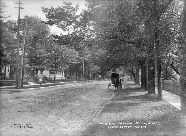 View along right side terrace, looking down tree-lined West Main Street. Houses are on the left side. Along the curb on the right a woman is standing beside a horse-drawn carriage. Caption reads: "West Main Street, Oconto, Wis."
