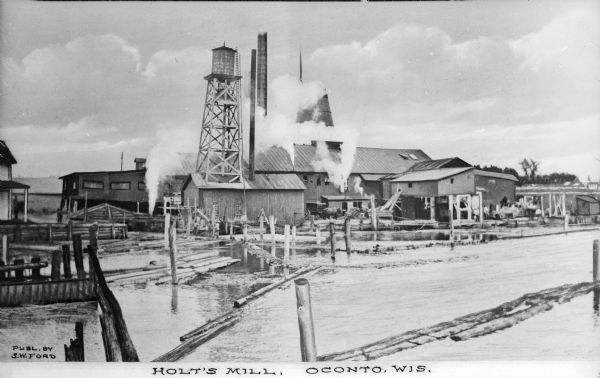 View across water toward the sawmill and various outbuildings located on the Oconto River. Steam is rising from a large pipe over over the main building, and from two smaller pipes beside it. Mechanisms in the river corral the raw logs to the structures for processing. Caption reads: "Holt's Mill, Oconto, Wis."