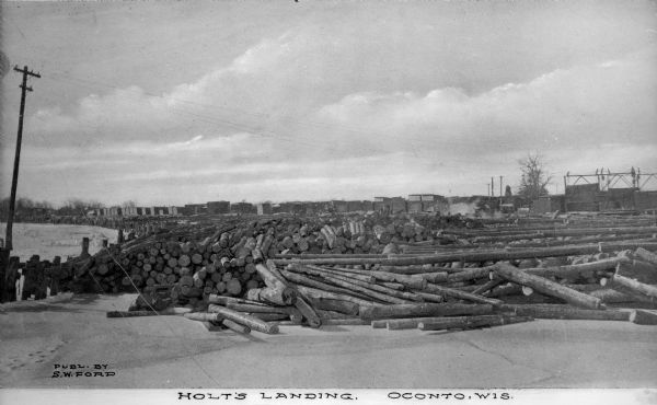 Large piles of logs waiting to be processed into lumber sit next to the Oconto River. In the background stand what appear to be piles of cut lumber. Caption reads: "Holt's Landing, Oconto, Wis."
