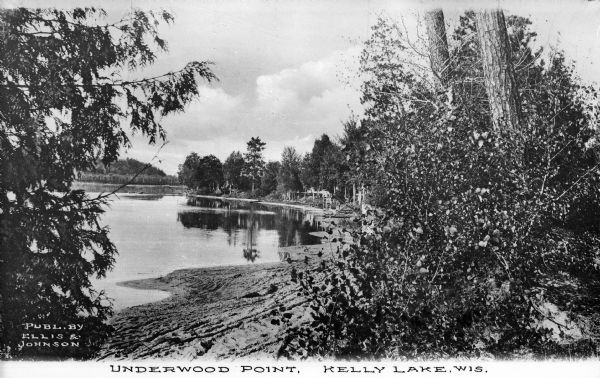 View of the tree-lined shore of Kelly Lake. Caption reads: "Underwood Point, Kelly Lake, Wis."