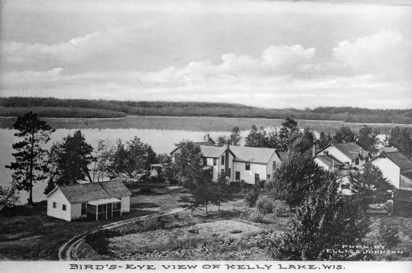 Elevated view of cottages along Kelly Lake's shoreline. Some of the buildings are two-story structures. Trees stand between the cottages and line the lake's opposite shore. Caption reads: "Bird's-Eye view of Kelly Lake, Wis."