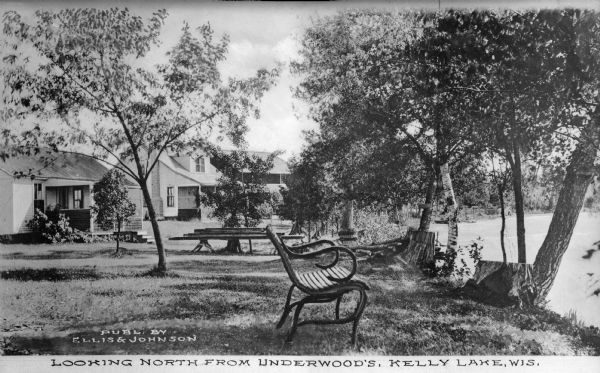View toward three cottages along the tree-lined shore of Kelly Lake at Underwood Point. Small docks with boats are along the shoreline. A wooden bench with scrolled arm rests faces the water in the foreground. Caption reads: "Looking north from Underwood's, Kelly Lake, Wis."