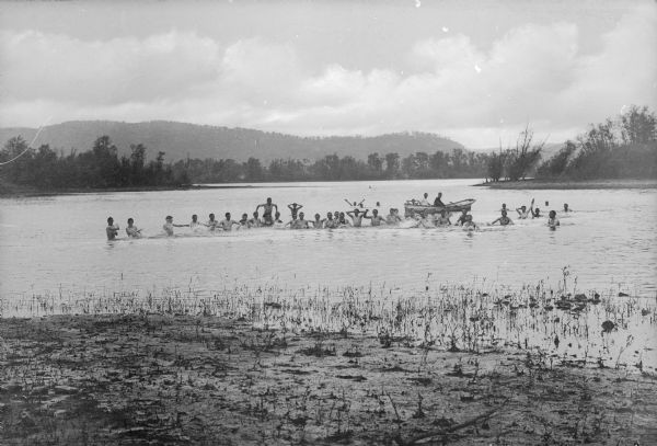 Boys swimming in a lake pose for a photograph as others sit in a rowboat behind them.
