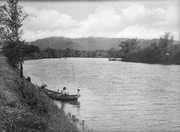 View down steep hill at the shoreline looking toward two young boys sitting in a rowboat below. A group of people in a boat are in the distance along the opposite shore. Hills are in the background.