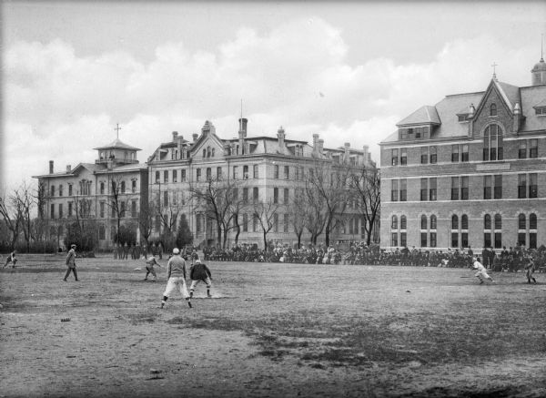 Boys play a baseball game while a large crowd of spectators watches from the sidelines in the background in front of three stone buildings. The building on the far left has a bell tower from which additional spectators watch the game.