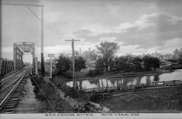 View along railroad tracks leading towards a railroad bridge across the Red Cedar River. A large building is on the opposite shoreline. Caption reads: "Red Cedar River, Rice Lake, Wis."
