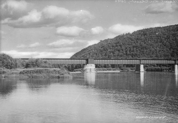 View across water toward a railroad bridge, constructed of heavy steel that sits on massive stone supports. Wooded hills are in the background.
