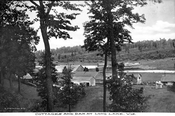 View down hill toward the dam and cottages at Long Lake. The structures are made of wood, with outbuildings in the foreground. Tree-covered hills are in the background. Caption reads: "Cottages and Dam at Long Lake, Wis."