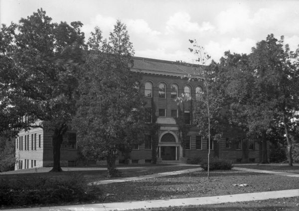 View across lawn toward the neoclassical-style facade of Ingraham Hall at Ripon College. The building is made of brick with a stone foundation. A column flanks each side of the double doors, and an arched window consisting of triangular panes sits above.