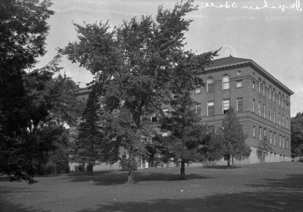 View looking uphill toward the back of Ingraham Hall at Ripon College. The primary material used is brick, and the foundation and basement are stone. The back entrance, placed on the same axis as the front entrance, mirrors the front, although the decoration around it is simpler. Half columns flank the the door, which is protected by an arched entryway. The third floor windows are arched.