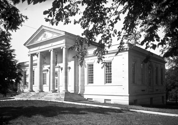 A view of the library's facade and side. This building follows the Neoclassical style popular in the early twentieth century, and it is an iconic style for public structures. The main entranced is above ground level, reached by a large set of steps, and is protected by a two-story porch, supported by columns, and above a pediment decorated with carvings.