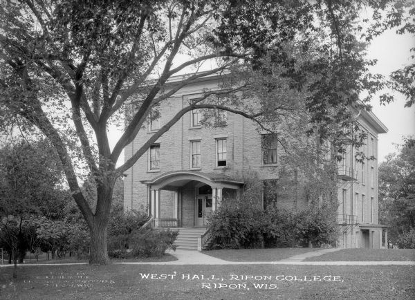 View across lawn toward the facade of West Hall at Ripon College. The entrance doors are elevated above ground level, reached by a large porch with curved roof. Caption reads: "West Hall, Ripon College, Ripon, Wis."