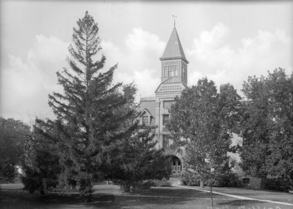 View across lawn toward the facade of Bartlett Hall at Ripon College, partially obscured by trees. Brick is the primary building material used, although large stones form the arch of the entrance. Ornamental brickwork is on the upper floors. A cupola forms the peak of the roof.
