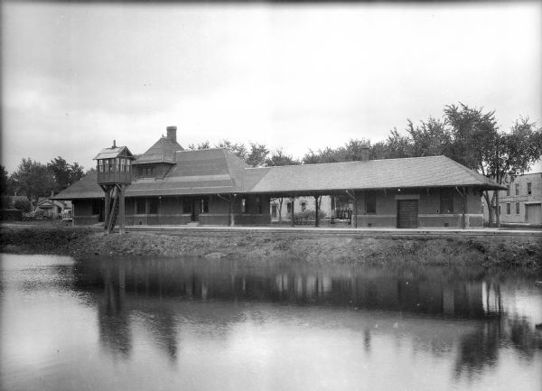 View across water toward the back side of a railroad station and outbuilding that face the creek. A hotel, garage, and homes are around the structure.