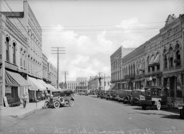 View down center of the downtown business district, with several people walking on the sidewalk past storefronts. Automobiles are parked at an angle along both sides of the street. The Grand View Hotel is in the distance.