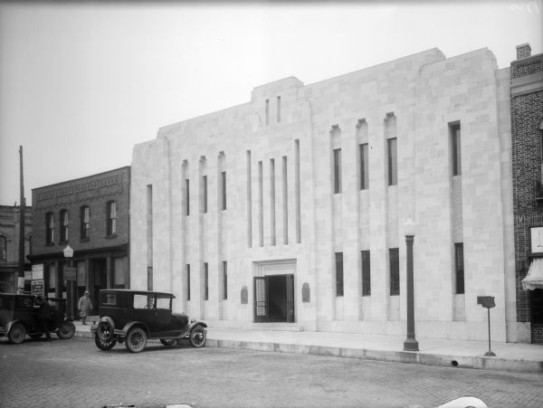 Two cars are parked in front of The First National Bank and a man walks down the sidewalk in front of the building. The bank is designed in an Art Deco style and features a stone facade.